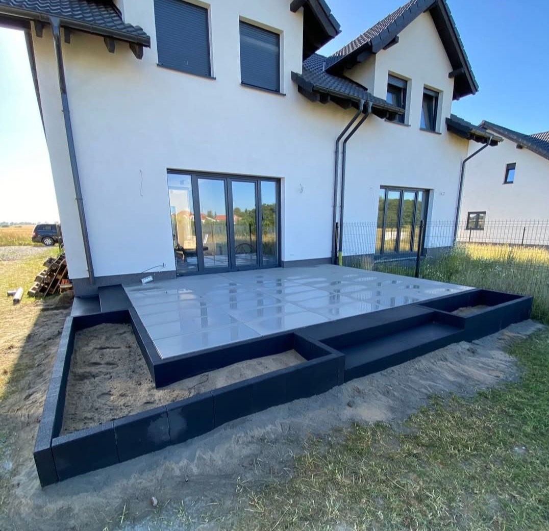 Nowoczesny taras z szarych płyt, otoczony czarną obudową z kostki. Widok na biały dom z szarymi oknami i ciemnym dachem w słoneczny dzień.