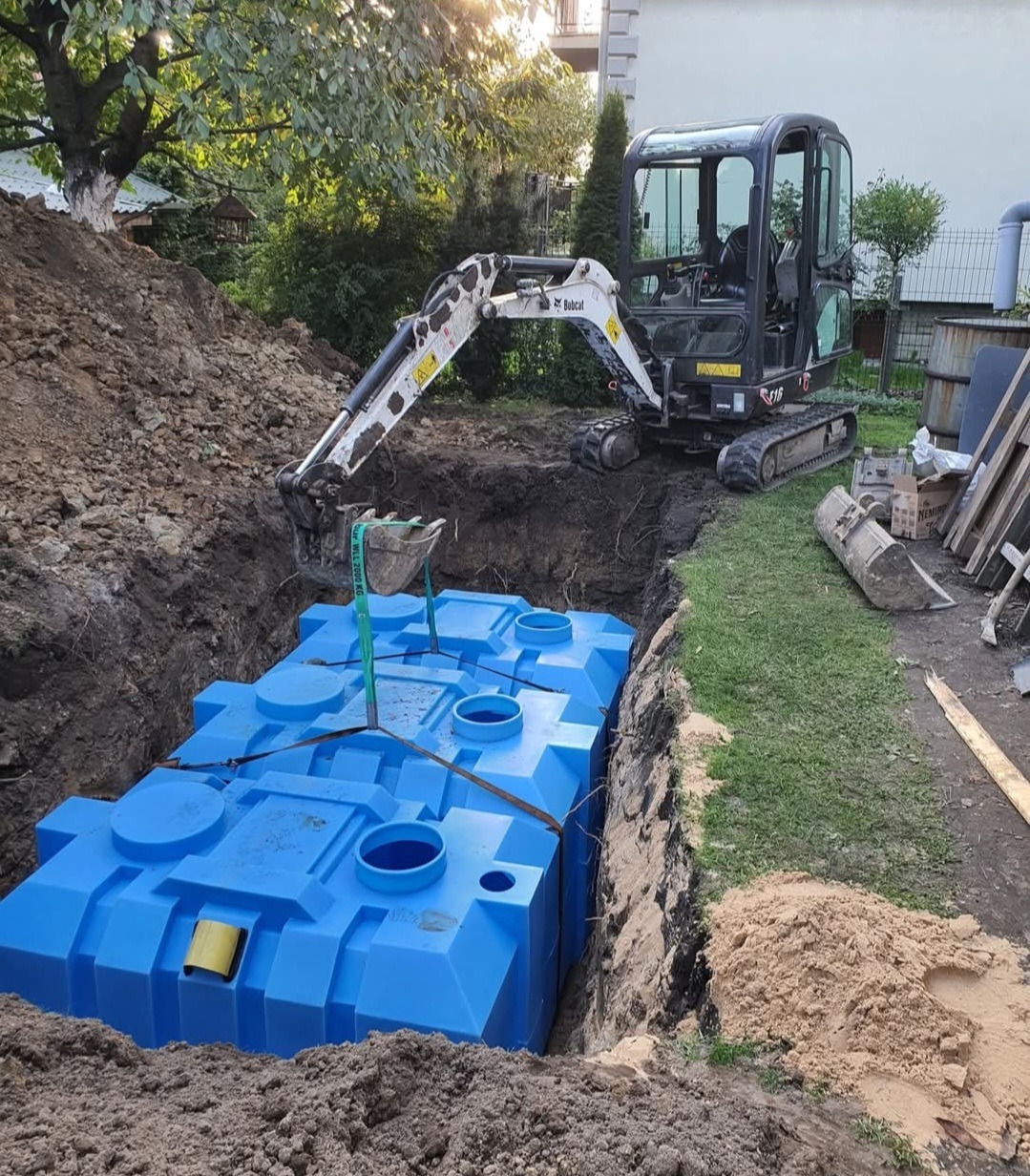 Montaż zbiornika retencyjnego z tworzywa sztucznego w wykopie, widoczny mały koparka. Etap instalacji, zielona trawa i drzewa w tle.