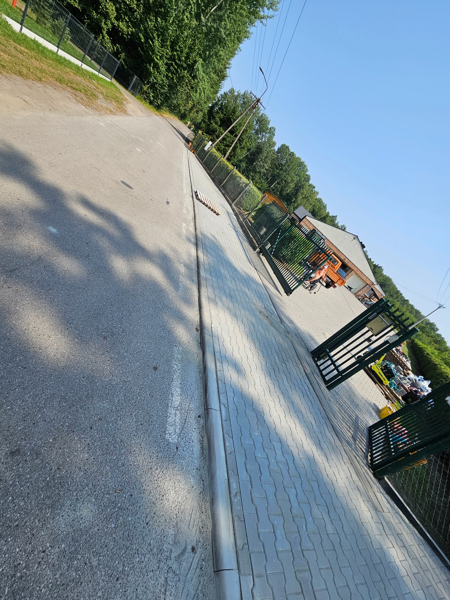 Otwarta, zielona brama wjazdowa z metalową siatką w tle, kostka brukowa, częściowo widoczny budynek i drzewa. Ujęcie z perspektywy ulicy.