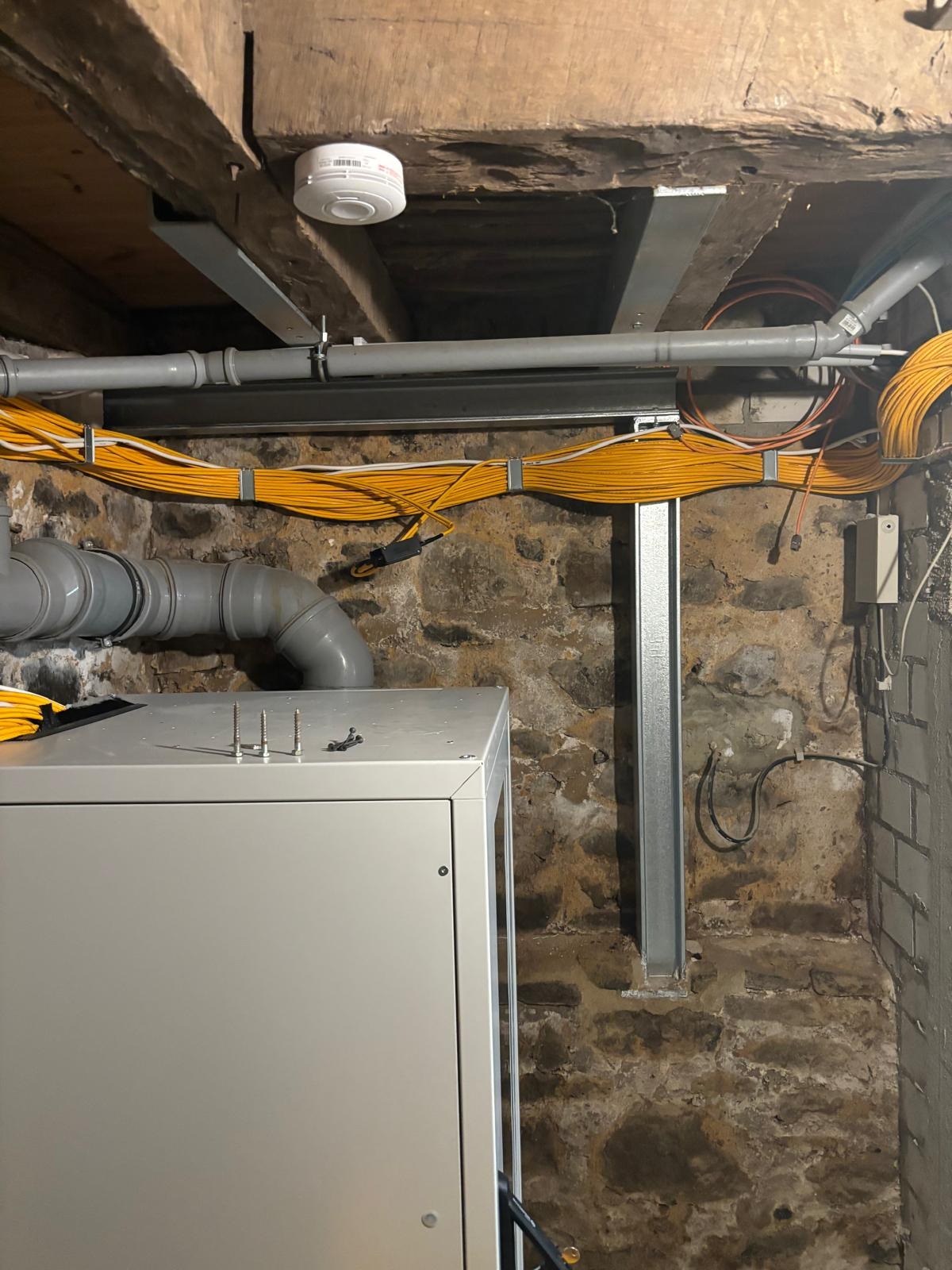 Instalacja elektryczna w piwnicy z rurami PCV i metalowymi wspornikami. Widoczne przewody w żółtej izolacji oraz elementy centrali. Kamienna ściana w tle.