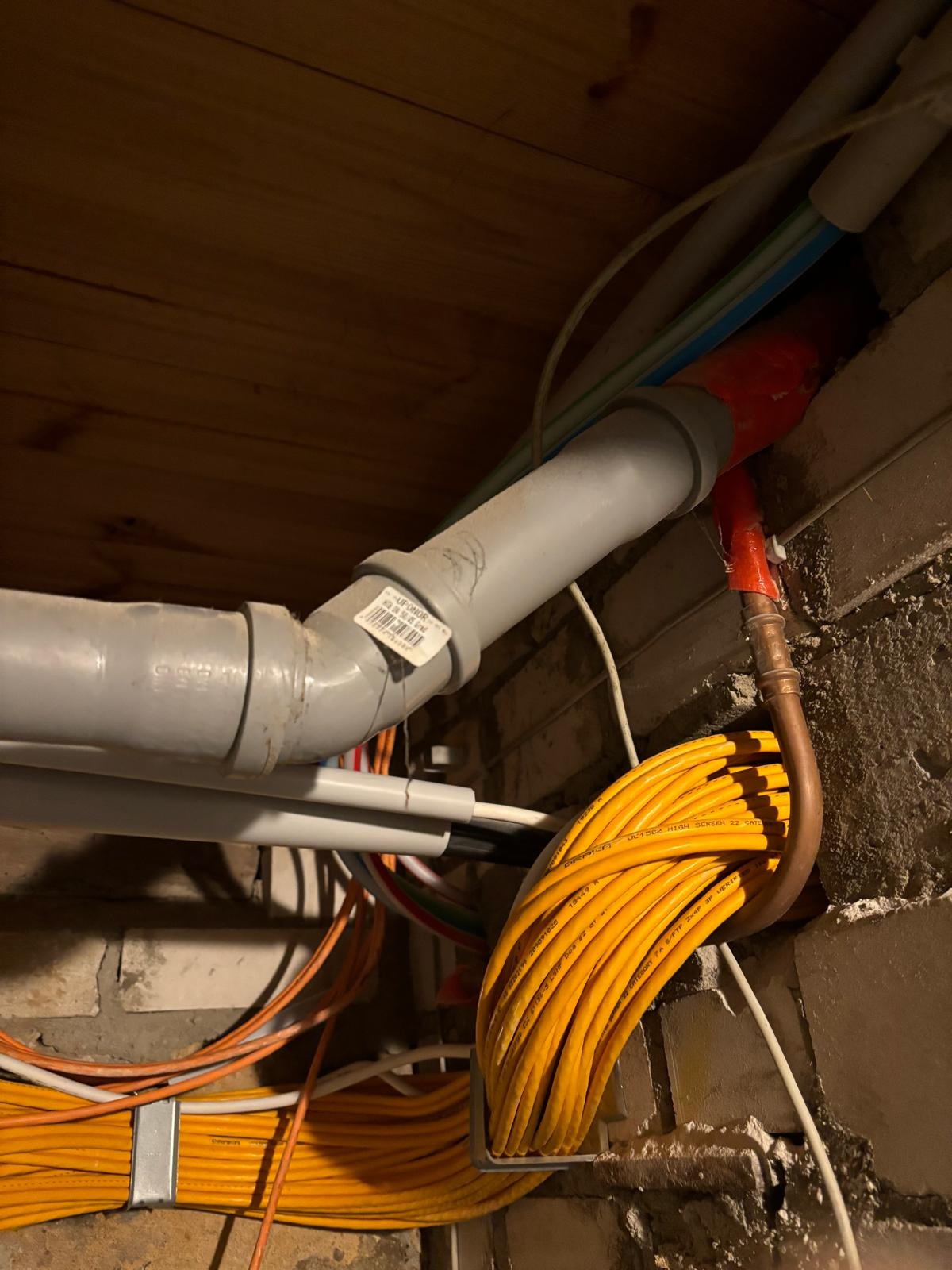Szare rury kanalizacyjne i wiązka żółtych kabli w surowym wnętrzu. Widoczne połączenia rur i mocowanie kabli do ściany z cegły. Fragment drewnianego sufitu.