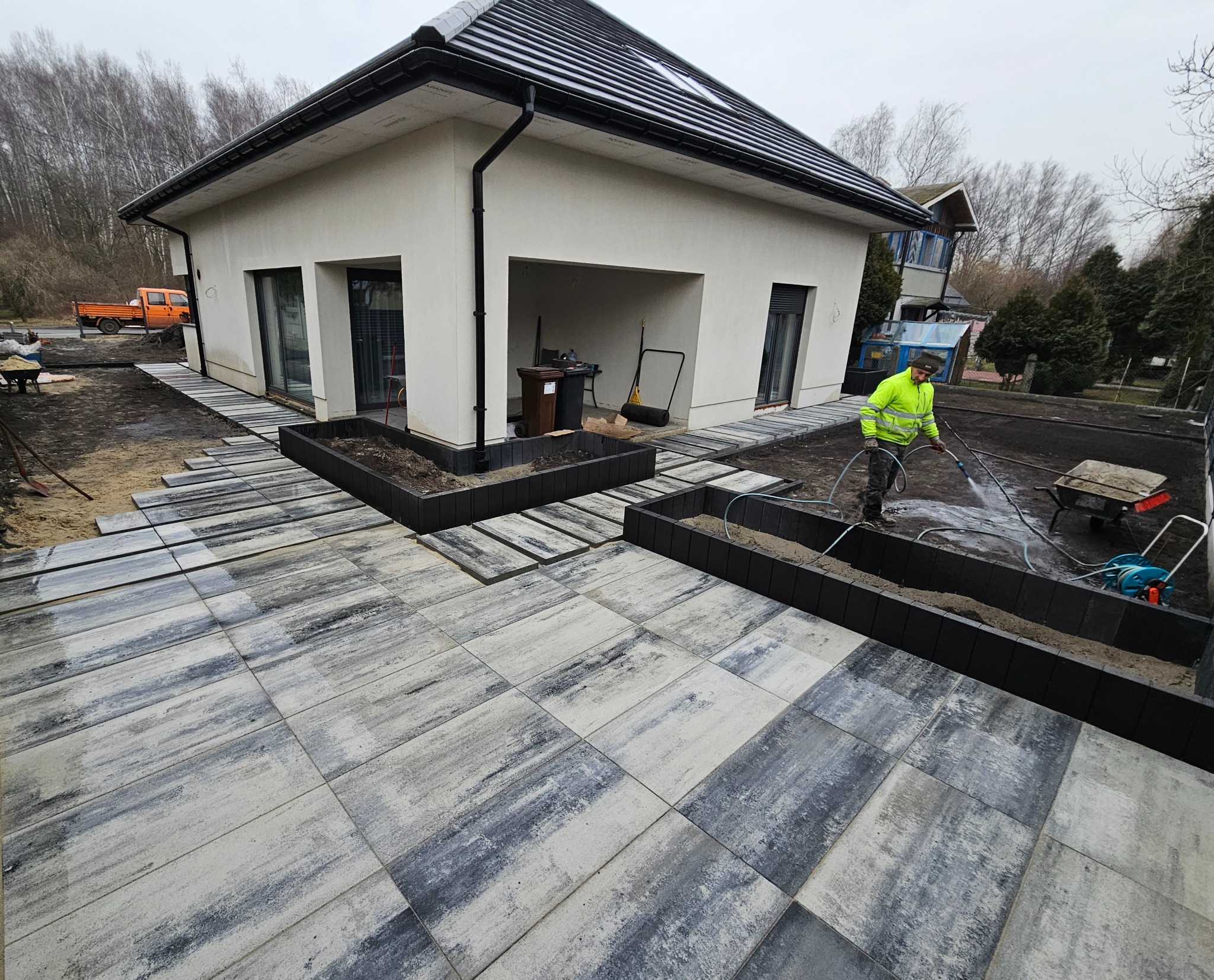 Układanie kostki brukowej na posesji przy domu. Pracownik w żółtej kurtce spryskuje powierzchnię wodą. Widoczne rabaty i elementy małej architektury.