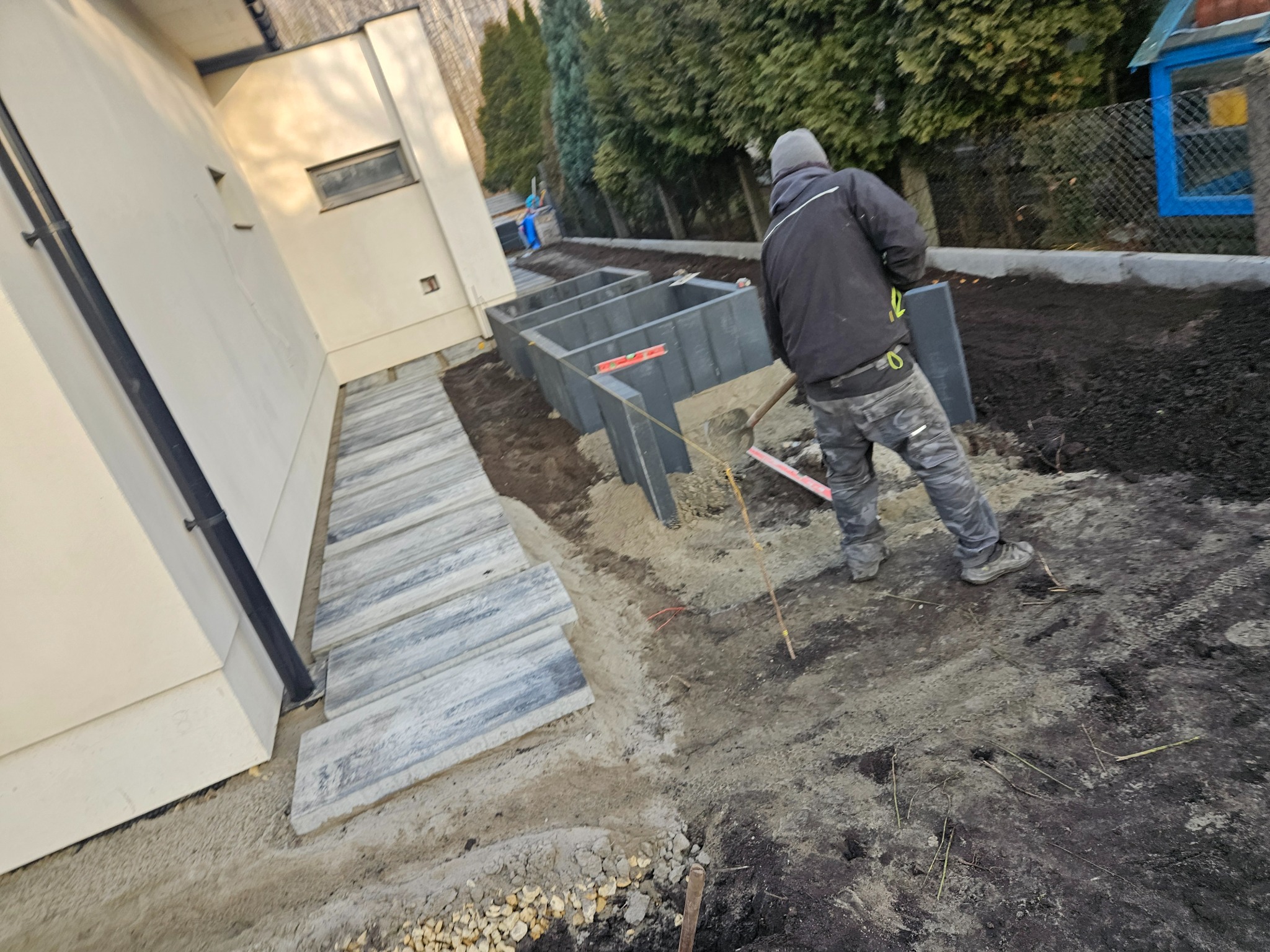 Układanie betonowych obrzeży rabat w ogrodzie. W tle widoczny pracownik z łopatą. Częściowo ułożony chodnik z płyt betonowych przy budynku.