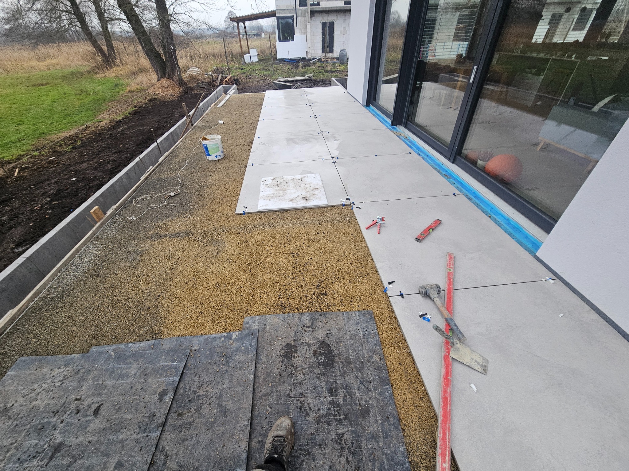 Układanie szarych płyt tarasowych na żwirowym podłożu przy nowoczesnym domu. Widoczne narzędzia: poziomica, kielnia i krzyżyki dystansowe. Prace w toku.