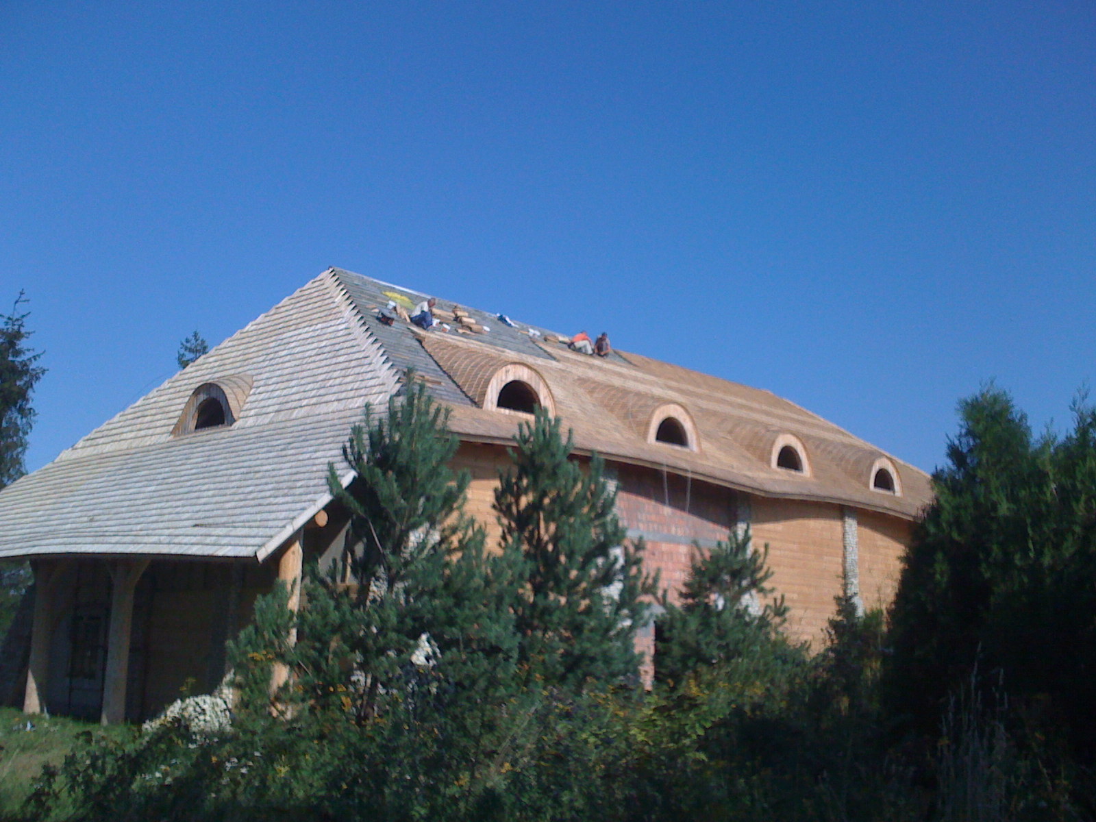 Budowa domu w stylu góralskim z drewnianą konstrukcją i dachem krytym gontem, widoczne okrągłe okna dachowe i ekipa dekarzy pracująca na dachu.