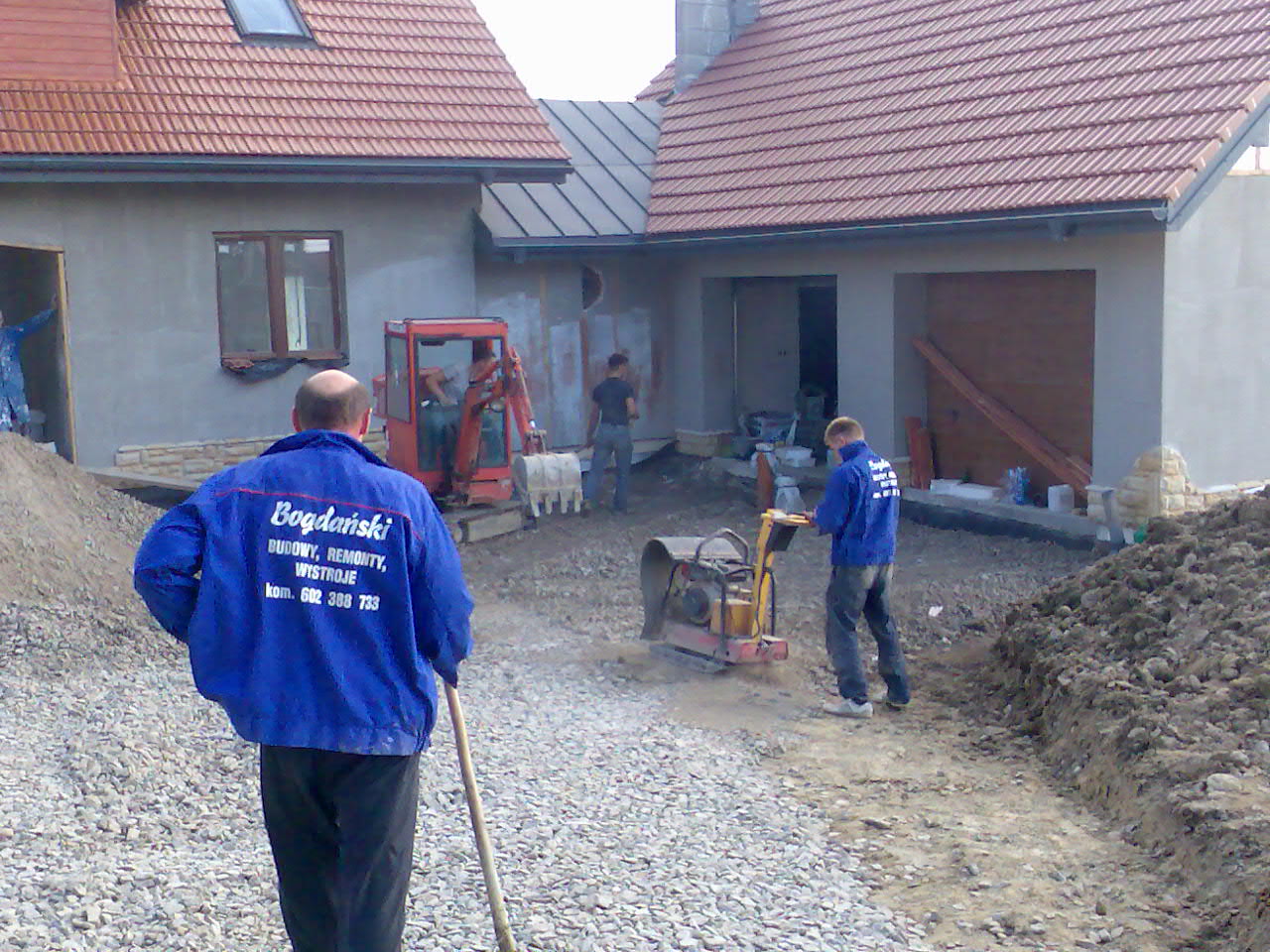 Prace ziemne przed budynkiem w stanie surowym; pracownik zagęszcza podłoże zagęszczarką wibracyjną, w tle minikoparka i pracownicy, na plecach jednego z pracowników widoczne logo firmy budowlanej.
