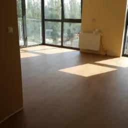 Jasne wnętrze z nową podłogą korkową, duże okna z widokiem na zieleń, grzejnik przy ścianie, promienie słoneczne na podłodze.