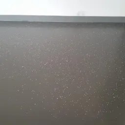 Szara, błyszcząca podłoga z drobnymi, jasnymi plamkami, widoczny fragment ściany i drzwi, listwa przypodłogowa w kolorze podłogi.