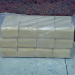 Pakiet jasnego brykietu drzewnego ułożonego w kostkę, zapakowany w przezroczystą folię, na tle płytek podłogowych i ściany w odcieniach czerwieni.