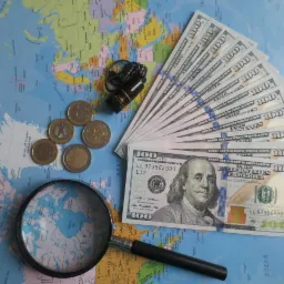 Mapa świata z lupą nad Ameryką Północną, obok euro monety, plik banknotów studolarowych oraz czarny dyktafon