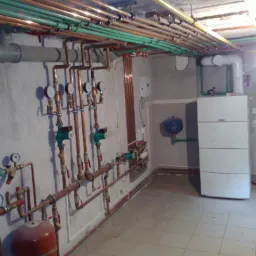 Piwnica z rozbudowaną instalacją hydrauliczną i grzewczą, widoczne rury miedziane, manometry, zawory, zbiornik wyrównawczy, piec grzewczy Vaillant i rury pod stropem.