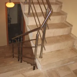 Marmurowe schody wewnętrzne z balustradą ze stali nierdzewnej, widok z góry na dół, z widocznym oświetleniem i otwartymi drzwiami w tle.