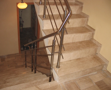 Marmurowe schody wewnętrzne z balustradą ze stali nierdzewnej, widok z góry na dół, z widocznym oświetleniem i otwartymi drzwiami w tle.