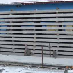 Stos prefabrykowanych betonowych płyt fundamentowych, ułożonych poziomo na metalowej konstrukcji, z widocznymi drewnianymi przekładkami i śniegiem na ziemi, na tle budynku z niebiesko-żółtą fasadą.