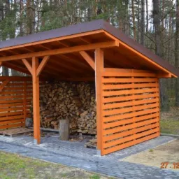 Drewniana wiata na drewno opałowe z brązowym dachem, z elementami ażurowymi, zbudowana na kostce brukowej, na tle lasu.