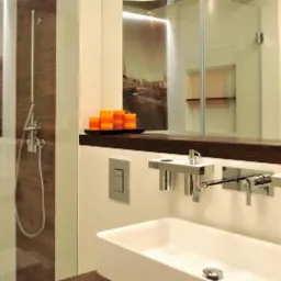 Nowoczesna łazienka z kabiną prysznicową, umywalką w kształcie prostokąta, chromowaną armaturą i dekoracyjnymi świecami w odcieniach pomarańczy, uzupełniona obrazem żaglówki.