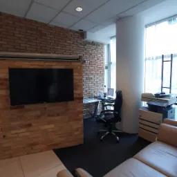 Nowoczesne biuro z ceglaną ścianą, dużym telewizorem, biurkiem z komputerem i beżową skórzaną sofą w pierwszym planie.