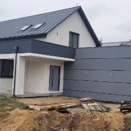 Nowoczesny dom jednorodzinny z szarą blachą na dachu i oknami dachowymi, widok z zewnątrz na plac budowy z piaskiem i deskami.