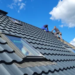 Montaż okien dachowych na ciemnoszarym dachu z dachówki ceramicznej, dwóch dekarzy w trakcie pracy na tle błękitnego nieba z chmurami.