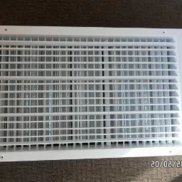 Biała kratka wentylacyjna z widocznymi otworami montażowymi, leżąca na brązowej powierzchni dywanowej, sfotografowana w jasnym świetle.