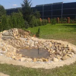 Oczko wodne z kamieni polnych i kaskadą z łupka, otoczone piaskiem, na tle zielonego trawnika i wysokich drzew iglastych.