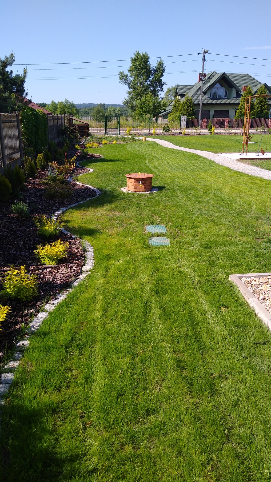 Zadbany trawnik z wyraźnymi pasami po koszeniu, z obrzeżami rabat wykończonymi kamieniem, studzienka z drewna i ścieżka z jasnego żwiru prowadząca do domu w tle.