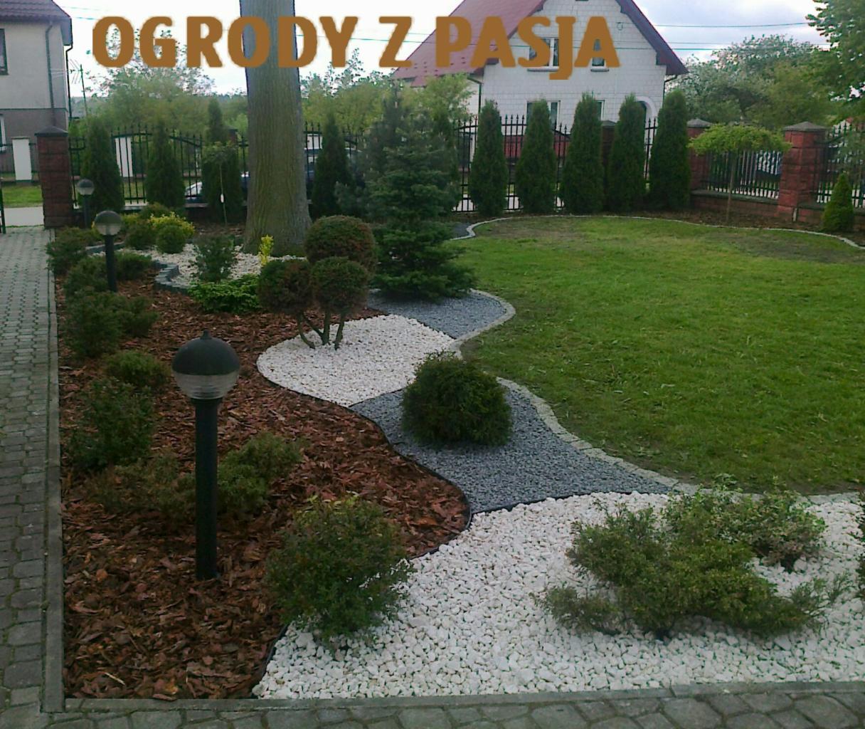 Ujęcie ogrodu z równo przystrzyżonym trawnikiem, ścieżką z białego i szarego żwiru, rabatami z kory i niskimi krzewami, z lampami ogrodowymi wzdłuż ścieżki i tujami w tle.