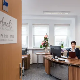 Wnętrze biura rachunkowego Aneks z widoczną pracownicą siedzącą przy biurku, komputerem i dokumentami, w tle kwiaty i okna z żaluzjami, na drzwiach tabliczka z godzinami otwarcia i certyfikatem...