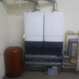 Dwa białe kotły grzewcze kaskadowe z brązowym zbiornikiem wyrównawczym, widoczne rury i instalacje w pomieszczeniu z kremowymi ścianami, instalacja połączona żółtymi rurami.