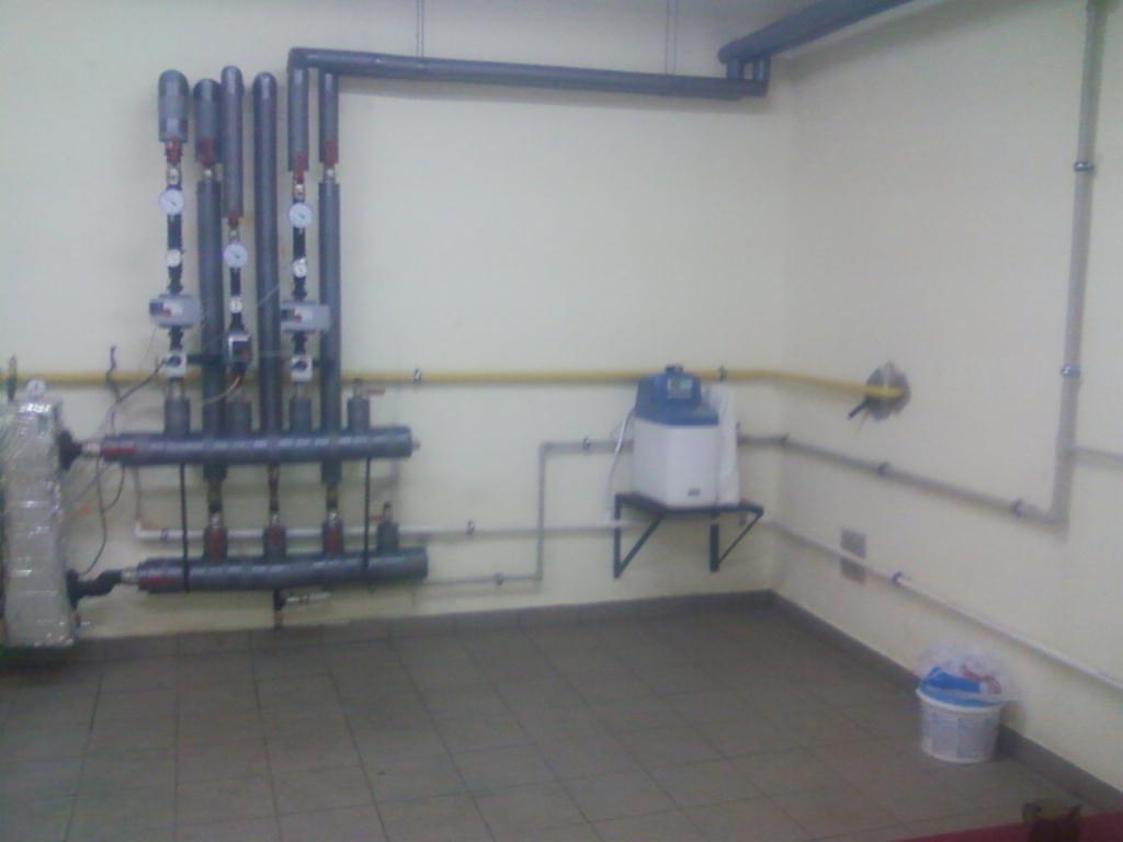 Kaskadowy system grzewczy z rurami i manometrami zamontowany na ścianie, widoczny zbiornik wyrównawczy i instalacja w pomieszczeniu technicznym.