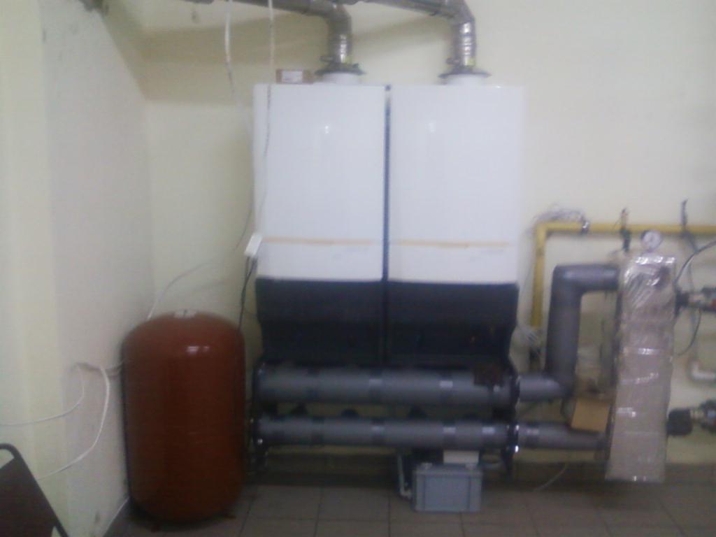 Dwa białe kotły grzewcze kaskadowe z brązowym zbiornikiem wyrównawczym, widoczne rury i instalacje w pomieszczeniu z kremowymi ścianami, instalacja połączona żółtymi rurami.