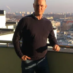 Mężczyzna w swetrze i jeansach stoi na balkonie z widokiem na miasto w oddali, z kominami fabrycznymi widocznymi na horyzoncie.