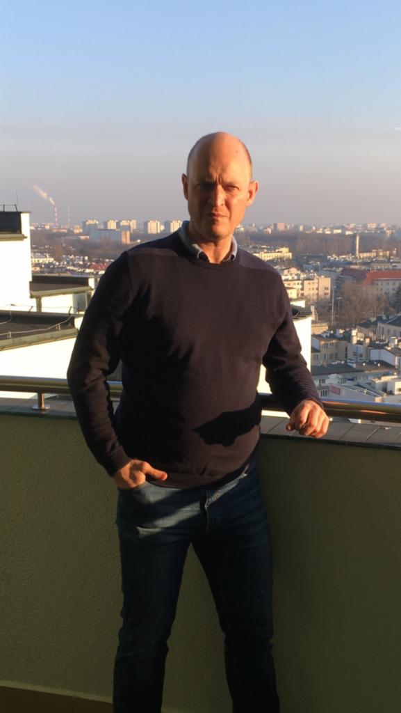 Mężczyzna w swetrze i jeansach stoi na balkonie z widokiem na miasto w oddali, z kominami fabrycznymi widocznymi na horyzoncie.