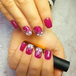 Dłonie z pomalowanymi na malinowy kolor paznokciami; na kilku paznokciach białe, koronkowe zdobienia. W jednej dłoni trzymana buteleczka lakieru.