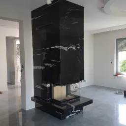 Nowoczesny, wysoki piec w obudowie z czarnego kamienia z białymi żyłkami, w surowym, jasnym wnętrzu.