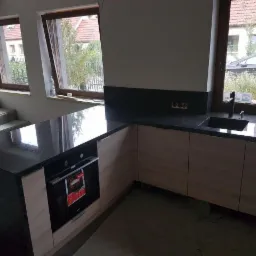 Nowoczesna kuchnia w trakcie wykończenia z blatem z ciemnego kamienia, jasnymi drewnianymi szafkami i wbudowanym piekarnikiem.