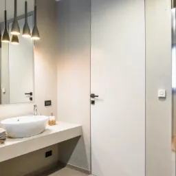 Nowoczesna łazienka z owalną umywalką na betonowym blacie, wiszące lampy nad lustrem, białe drzwi i kabina prysznicowa wyłożona mozaiką.