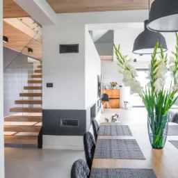 Jasne wnętrze z drewnianymi schodami z przeszkloną balustradą, dużym wazonem z białymi mieczykami na drewnianym stole z szarymi podkładkami i krzesłami oraz szarą sofą w tle