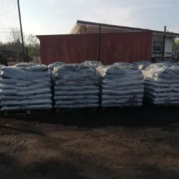 Rzędy worków z opałem, owiniętych folią, ułożonych na paletach na ziemi, z budynkiem magazynowym w tle.