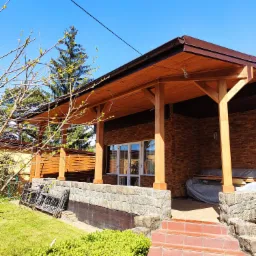 Nowoczesny dom murowany z drewnianym zadaszeniem nad wejściem, kamienną elewacją i schodami wyłożonymi czerwoną terakotą, widoczne rusztowania i materiały budowlane pod zadaszeniem.