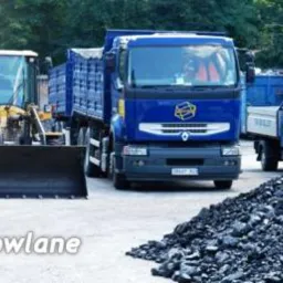 Kopalnia węgla: żółta ładowarka, niebieska ciężarówka i hałda węgla kamiennego na placu składowym. W tle kontenery i drzewa.