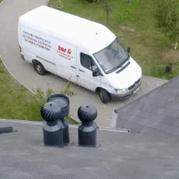 Dwa turbowentylatory na dachu budynku, w tle biały bus firmy instalacyjnej na tle zieleni. Widok z góry.