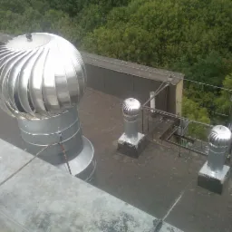 Metalowe turbowentylatory na dachu budynku z widokiem na zieleń. Jeden turbowentylator jest większy od pozostałych dwóch. Widok z góry, lekko z boku.