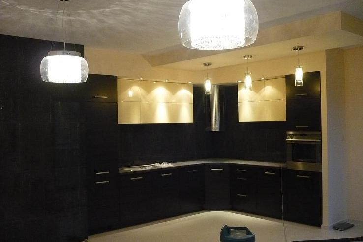 Nowoczesna kuchnia z ciemnymi, lakierowanymi szafkami dolnymi i jasnymi szafkami górnymi podświetlonymi od spodu, wiszące lampy z okrągłymi kloszami, widoczny fragment podłogi i pojemnik na środki...