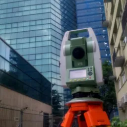 Tachimetr na pomarańczowym statywie ustawiony na chodniku, w tle nowoczesny szklany biurowiec i budynek w rusztowaniach.