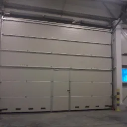 Zamknięta, segmentowa brama przemysłowa w hali magazynowej, z widocznymi prowadnicami i konstrukcją nośną. Fragment okna w tle. Brama w jasnym odcieniu szarości.