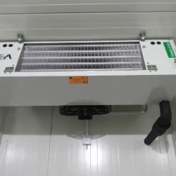 Widok z dołu na zamontowany pod sufitem chłodniczy agregat przemysłowy marki GEA Kuba Green Line z widocznym wentylatorem i odpływem skroplin w pomieszczeniu z panelowych ścian.