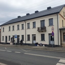 P.H.U. MAJSTER-BUD Jacek Kozłowski - Pracownicy przy białej elewacji budynku, jeden na drabinie, inni na ziemi. Widoczny rowerzysta i znak przystanku. Czarny dach i okna. Ulica z liniami.