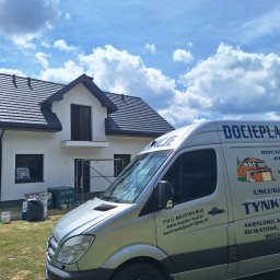 P.H.U. MAJSTER-BUD Jacek Kozłowski - Nowoczesny dom z białą elewacją i czarnym dachem, widoczny zza srebrnego busa z reklamą firmy budowlanej. Na trawniku stoją puszki z farbą i pracownik.