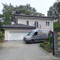 P.H.U. MAJSTER-BUD Jacek Kozłowski - Jasny, dwukondygnacyjny dom z garażem i balkonem, po renowacji elewacji. Na podjeździe zaparkowany srebrny bus z reklamą usług budowlanych. Widok z zewnątrz, w tle drzewa.